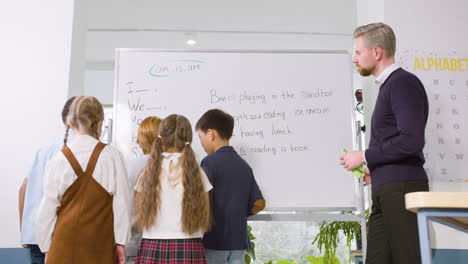 Rückansicht-Einer-Multiethnischen-Schülergruppe-Und-Eines-Lehrers,-Die-Vor-Der-Tafel-Stehen-Und-Das-Verb-„to-Be“-Studieren