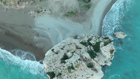 an aerial view shows the famous aphrodite's rock, a sea stack in paphos, cyprus, with waves lapping around it and a sandy beach nearby