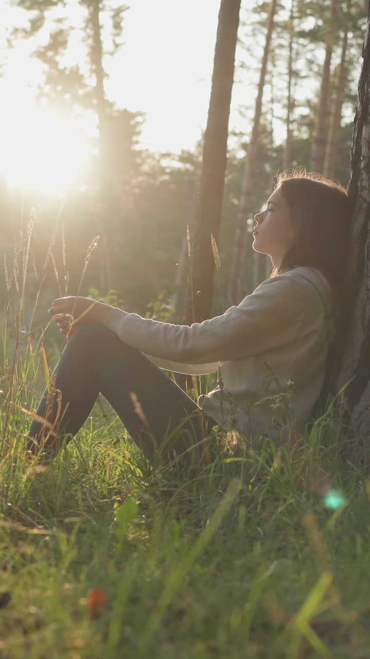 dama se apoya en la corteza del tronco del árbol en el bosque al atardecer. mujer joven descansa en el aire fresco pensando en la mejora de la salud mental. lidiar con problemas personales en la naturaleza