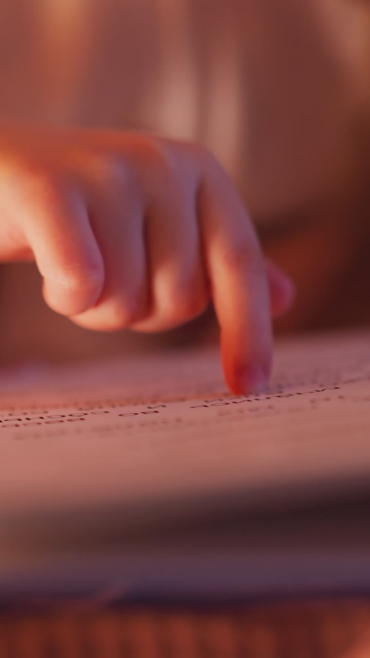 Hand of little kid follows line on page with finger while girl reads book covered with warm blanket in semidark bedroom extreme close view slow motion