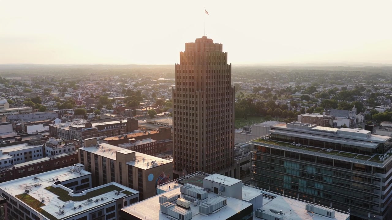 Centro de Allentown, Pensilvânia, ao pôr do sol, com torres de escritórios modernas, bairros residenciais e luz quente do entardecer. Plano geral. Tomada aérea de aproximação por drone. Bandeira patriótica no topo