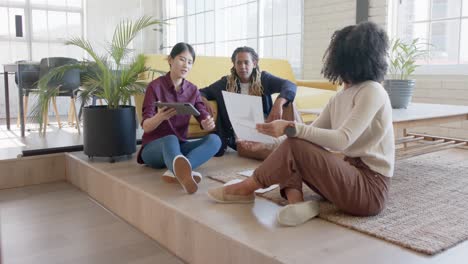 Happy-diverse-creative-colleagues-using-tablet-and-discussing-sitting-on-office-floor,-slow-motion