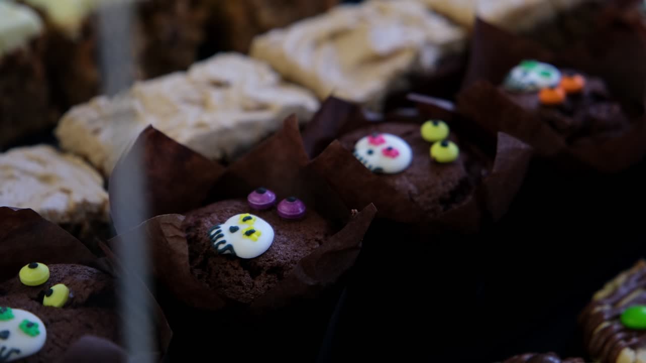 A close-up of chocolate cupcakes decorated with small skull candies and colourful toppings, surrounded by iced dessert bars on a tray. Perfect for Halloween, festive baking, or dessert themes
