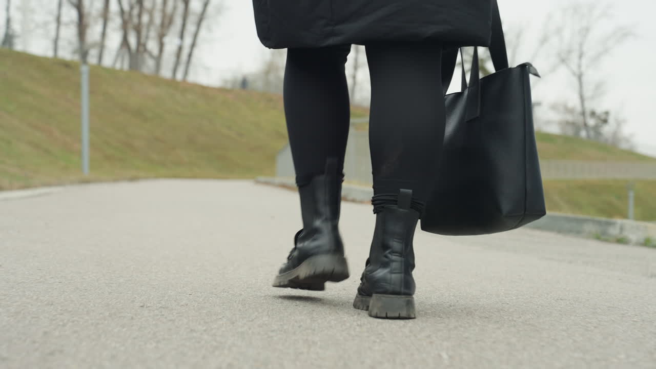Paso rápido con abrigo de invierno avanzando. Un bolso tote negro al costado mientras las botas recorren el pavimento. Una persona vestida con abrigo de invierno camina con confianza por una calle suburbana con su bolso y pasos firmes.