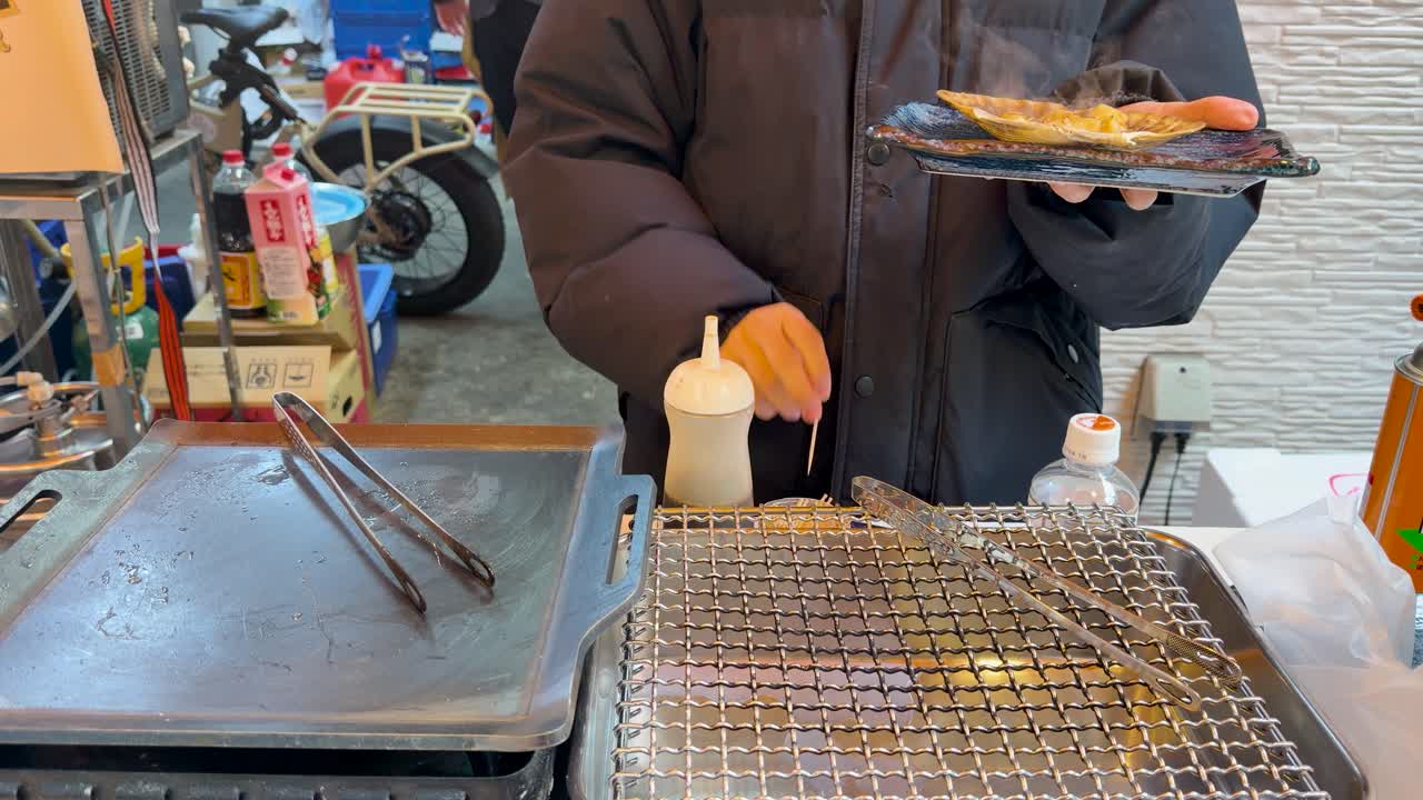 日本の市場の屋台で、店主がバター醤油で焼いたホタテを調理している。