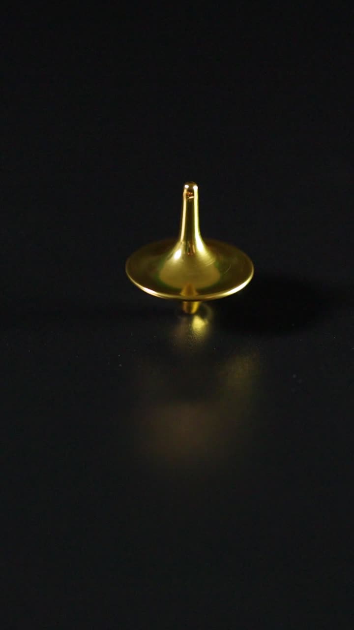Um pião de metal dourado gira suavemente em uma superfície escura e reflexiva, em orientação vertical.
