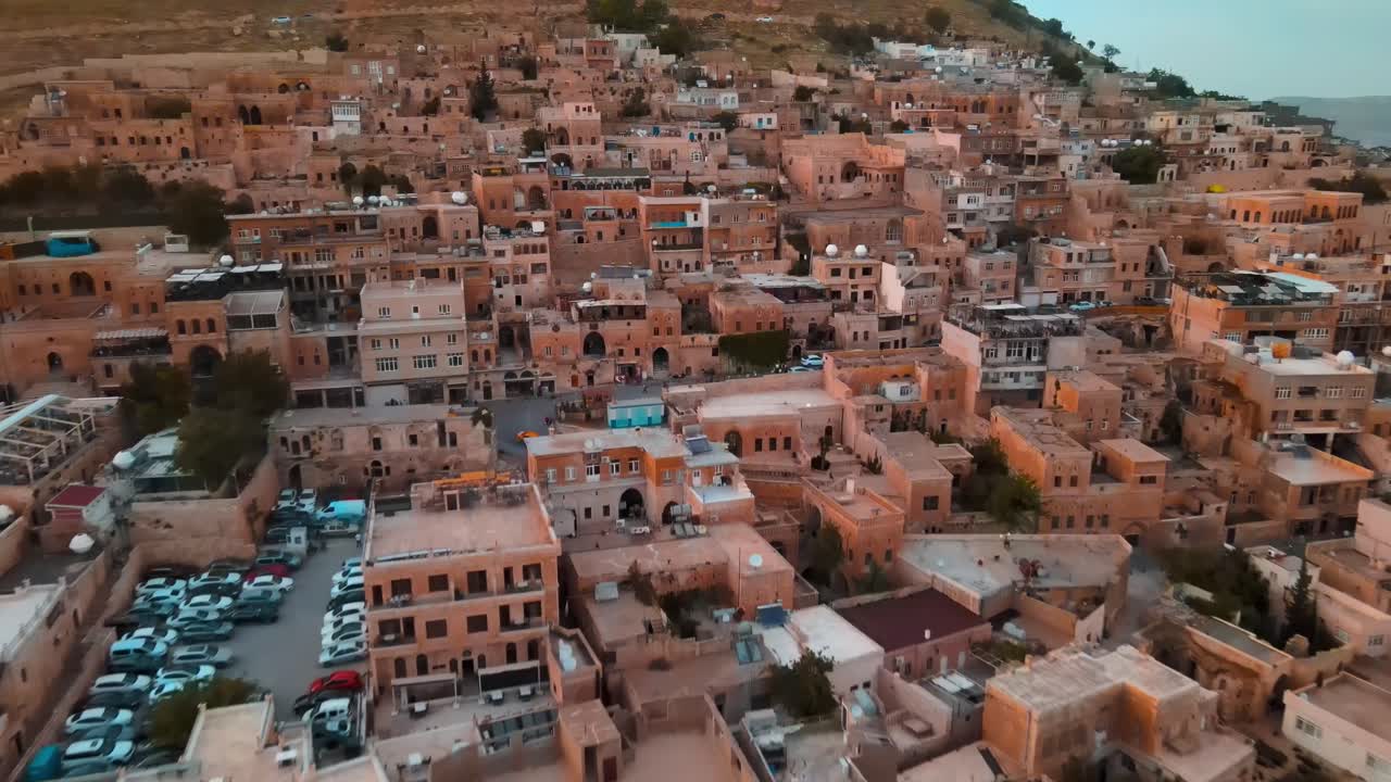 トルコのマルディン旧市街の夕暮れのドローン空撮。丘陵に石造りの建物が密集し、温かい夕暮れの色合いが広がる。歴史的な都市景観と文化的な観光地。