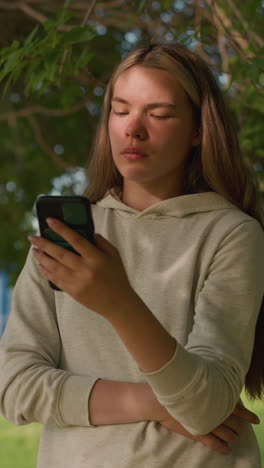 woman stands under tree with phone, deeply focused on screen, hand folded, while sunlight filters through leaves casting soft shadows on her face, background includes green scenery and a building