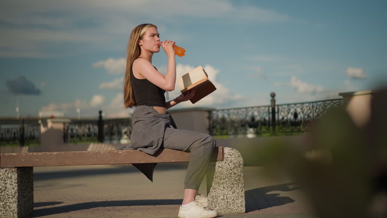 Woman seated outdoors, holding book in one hand while sipping juice from a bottle with the other, continuing to read, her shadow is cast on the ground, with blurred figures moving in the distance