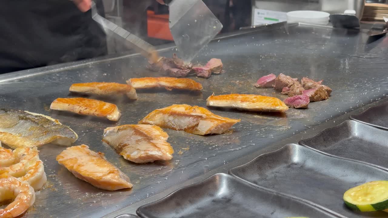 Close-up of a teppanyaki chef’s hands holding stainless steel spatulas, flipping golden, sizzling salmon on the cooktop. Steam rises from the grill.