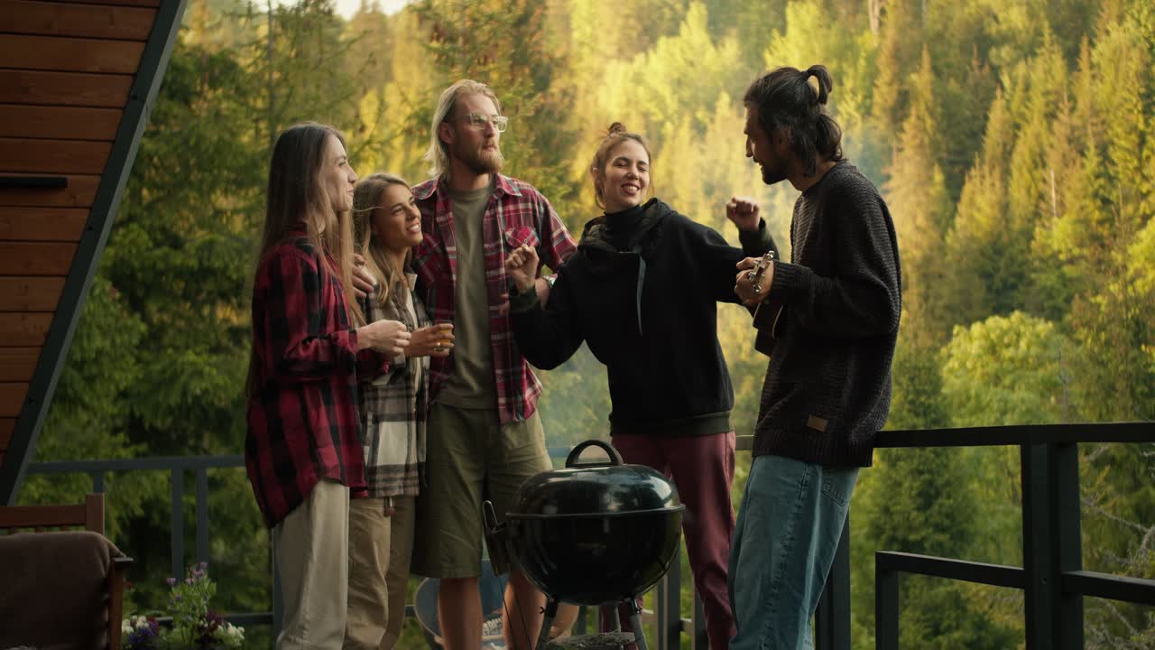 atmósfera cálida en el picnic: el tipo toca la guitarra y sus amigos bailan. entretenimiento mientras cocina en la parrilla contra el fondo de las montañas y el bosque de coníferas