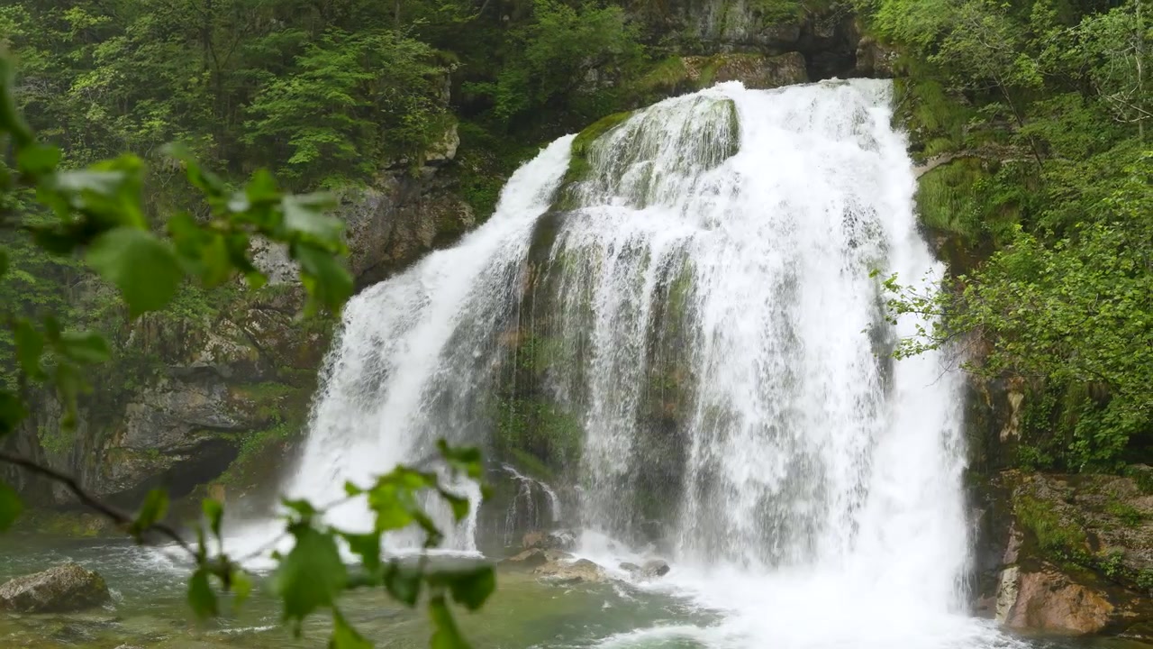 Vista primaverile della cascata Slap Virje attraverso le foglie in Slovenia, con un potente flusso di scioglimento della neve attraverso una foresta lussureggiante.