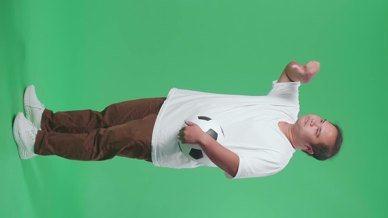푸른 스크린 배경에서 축구를 응원하는 동안 패자들을 조롱하는 몸짓을 보여주는 엄지손가락을 아래로 보여주는 공을 가진 아시아 남자의 몸이 가득 차 있습니다.