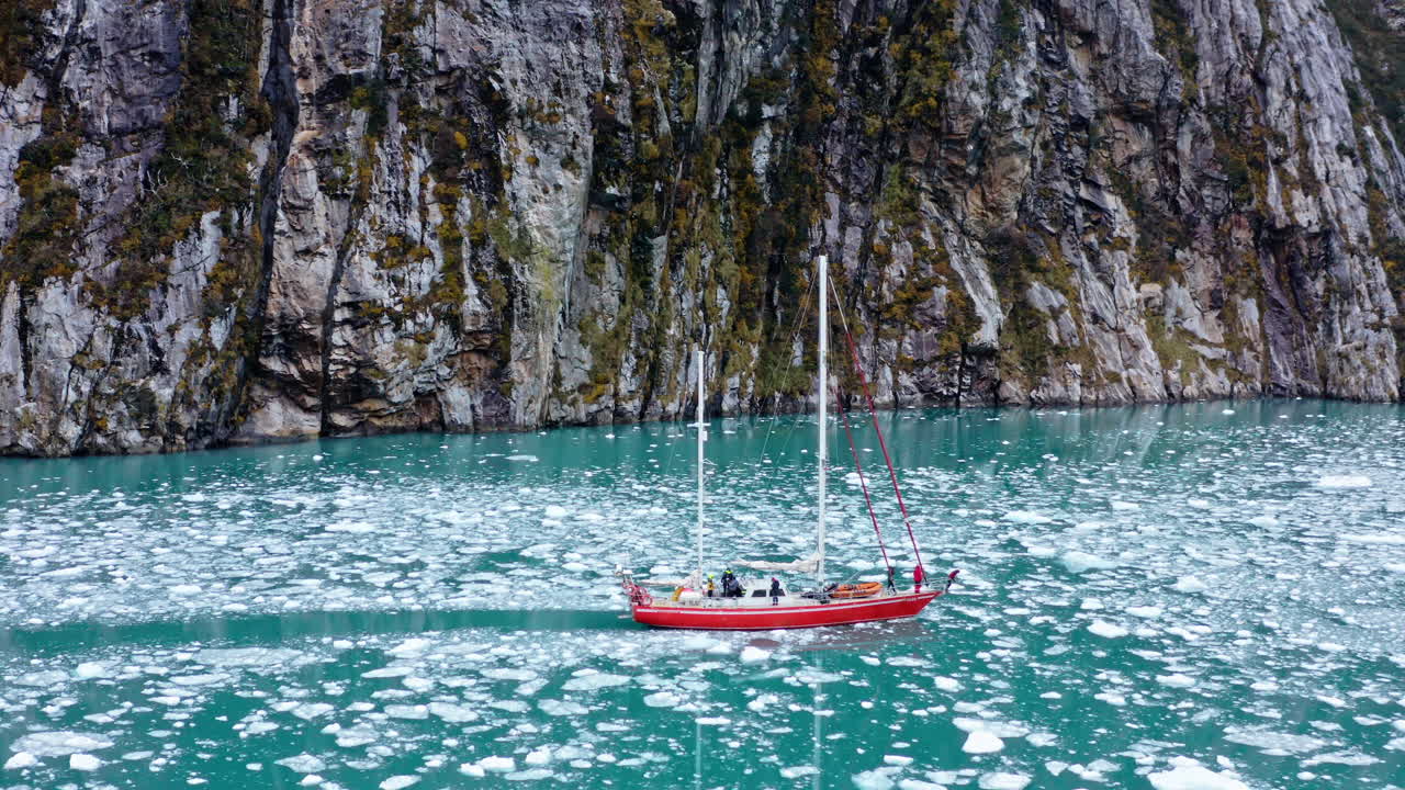Red sailboat cruises past steep rocky walls in calm clear icy water in remote Patagonian fjord, aerial orbit