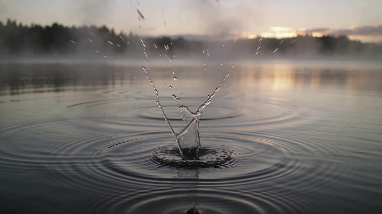 Recording falling droplet creating tall splash column and broad ripples on mist lake, with treeline