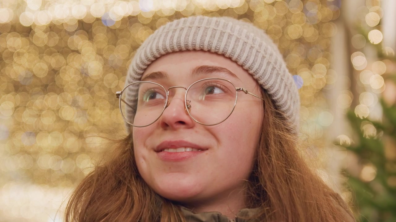 Hvid kvinde smiler under gyldne lys, med hue og briller, bløde lukkede øjne, et mildt udtryk af taknemmelighed, juletræ-bokeh, hyggelig jakke, fredelig aftenstemning, reflekterende varme.