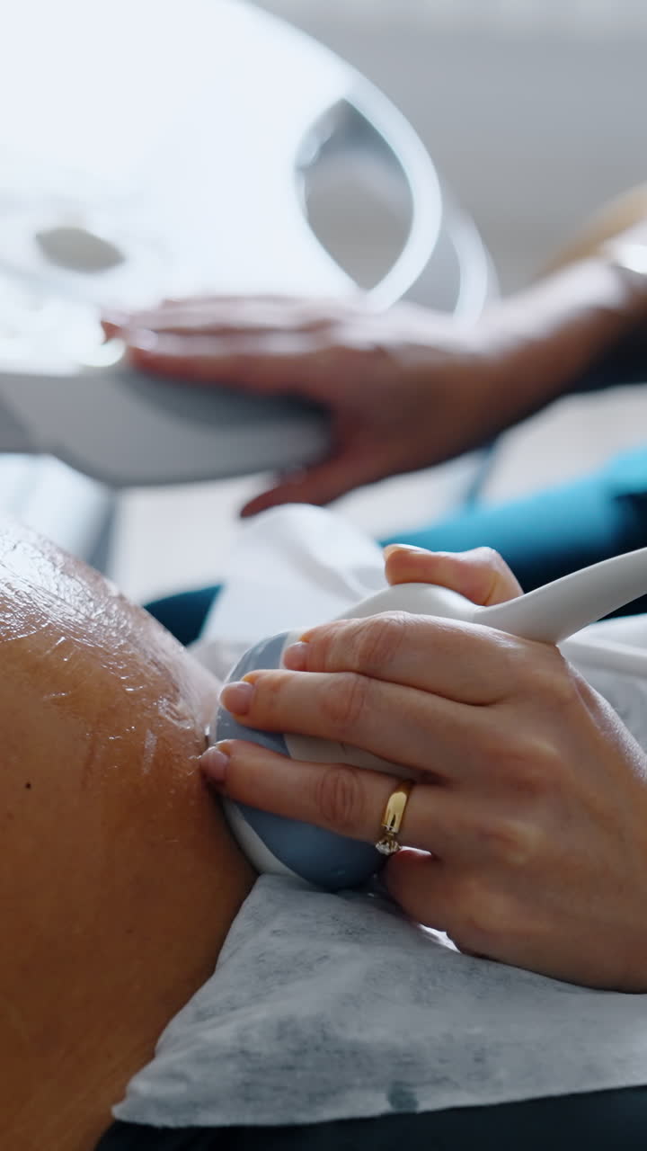 Cropped image of a pregnant lady having ultrasound check up. Hand of medic holds the device on belly and another hand operates ultrasonic machine. Close up.. Vertical video