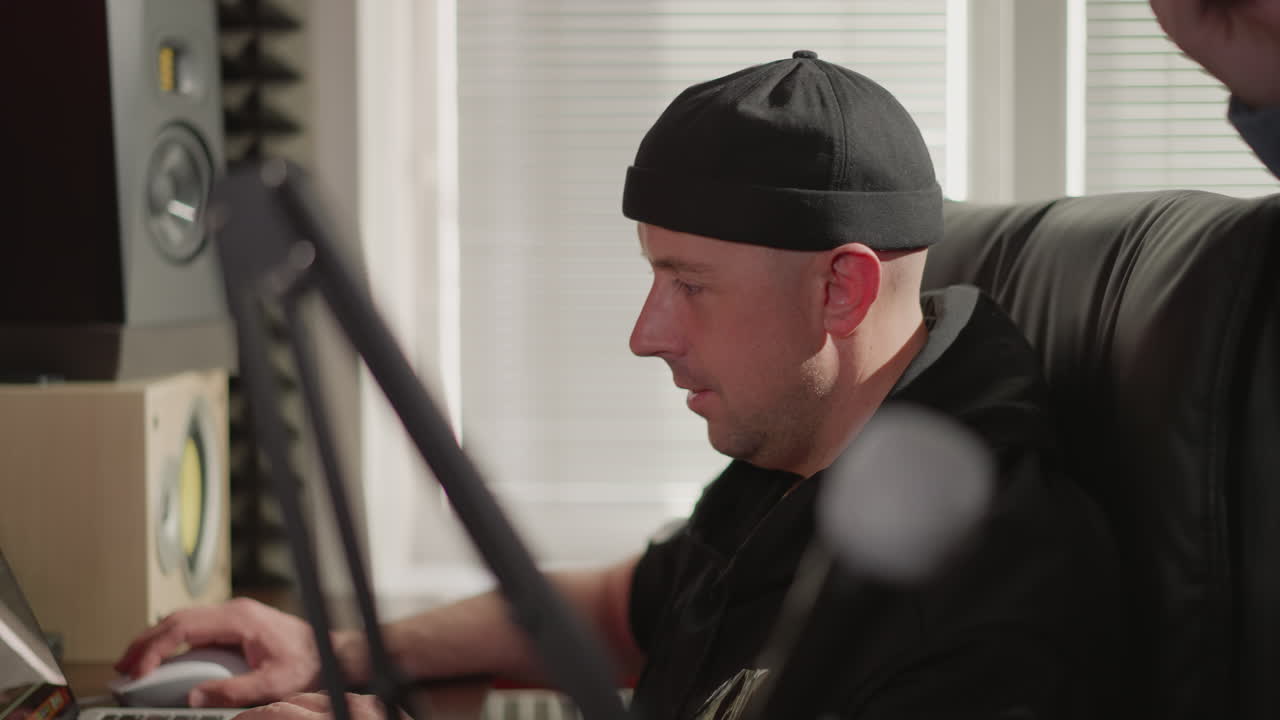 A man dressed in black, focused on working at a computer in a recording studio, sits at a desk surrounded by sound equipment. The setting includes studio monitors and acoustic foam panels