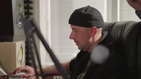 a man dressed in black, focused on working at a computer in a recording studio, sits at a desk surrounded by sound equipment. the setting includes studio monitors and acoustic foam panels