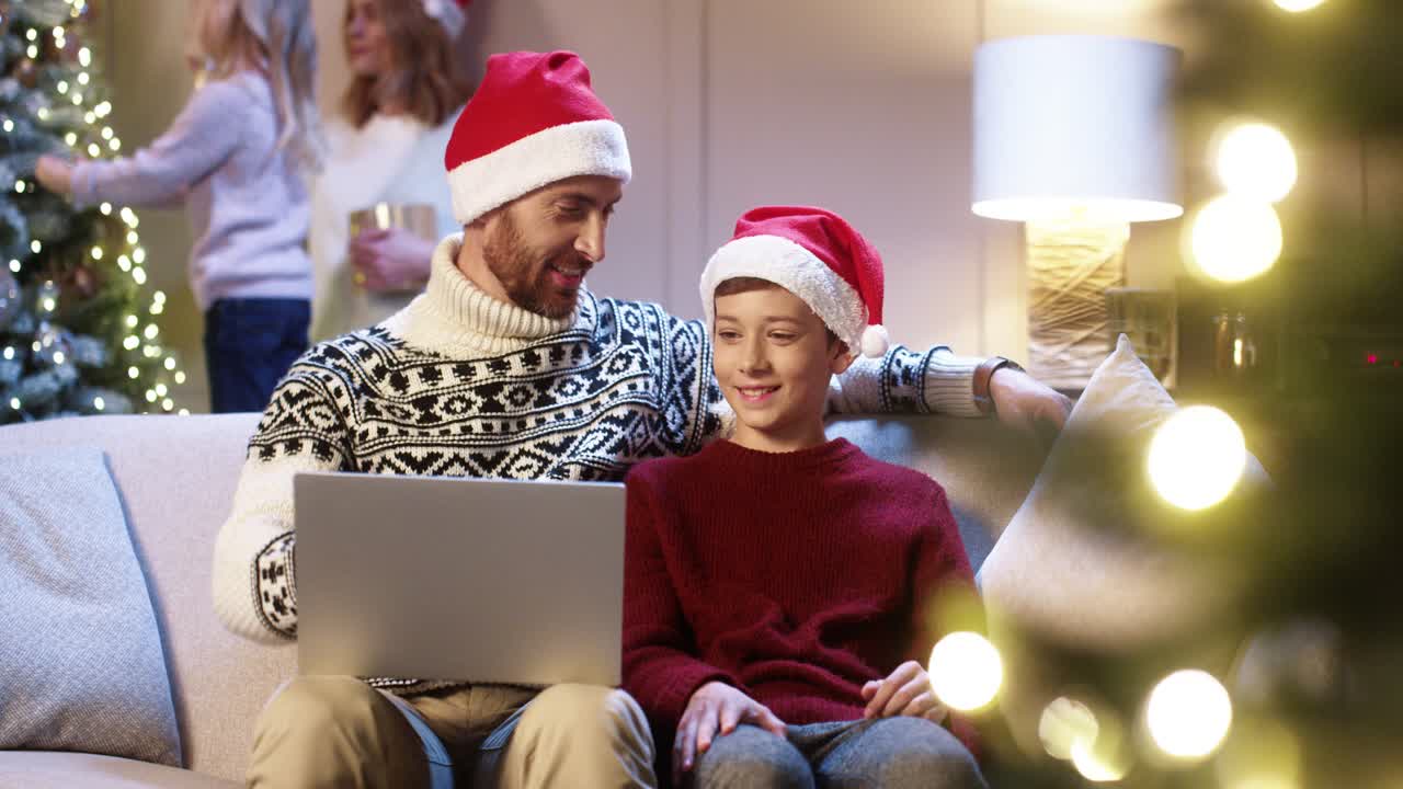 padre feliz con sombrero de santa con un hijo adolescente sentado cerca de un árbol de navidad decorado y brillante escribiendo en una laptop pasando las vacaciones de invierno en internet comprando regalos