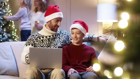 happy dad in santa hat with teen son sitting near glowing decorated christmas tree typing on laptop spending winter holiday time on internet buying gifts