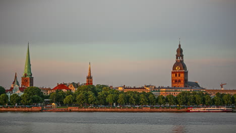 Time-lapse-shot-of-cruising-boats-on-river-and-cars-on-coastal-road-in-Riga-during-sunset-light---Famous-Cathedral-and-Tower-in-background