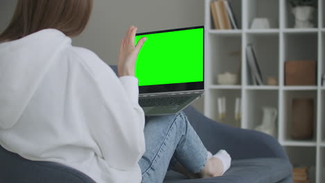 woman at home sitting on a couch works on a laptop computer with green mock-up screen. coronavirus covid-19 quarantine remote education or working concept. girl using computer browsing through