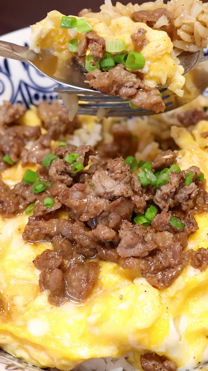 Uma sequência em close-up mostra uma pessoa usando uma colher e um garfo para comer carne moída, ovo mexido e arroz em uma tigela estampada sob iluminação brilhante.