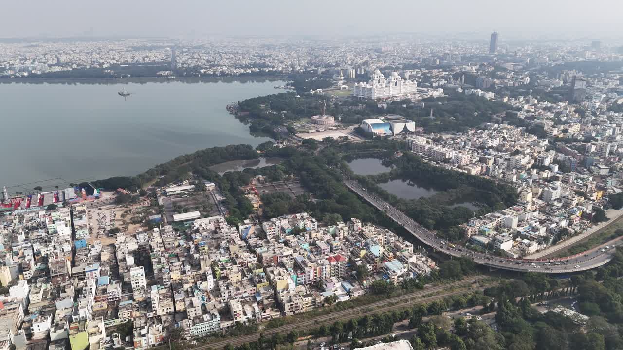 Ampia vista aerea di Hyderabad con Hussain Sagar, quartieri urbani densi e punti di riferimento iconici sotto un cielo diurno velato. Le strade Telugu Thalli Flyover e NTR Marg sono percorse da veicoli