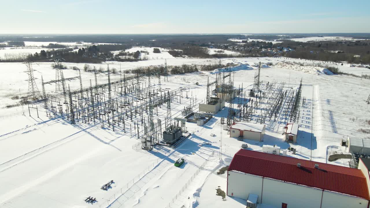 Aerial lateral view tracks across electrical substation with transformers, transmission pylons powering digital infrastructure in remote snowy winter fields. Energy network in cold winter conditions