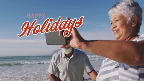 Animation-Von-Weihnachtsgrüßen-über-Ein-älteres,-Vielfältiges-Paar,-Das-Am-Strand-Ein-Selfie-Macht