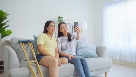 two women taking a selfie on a sofa. one has crutches and the other has a prosthetic leg.