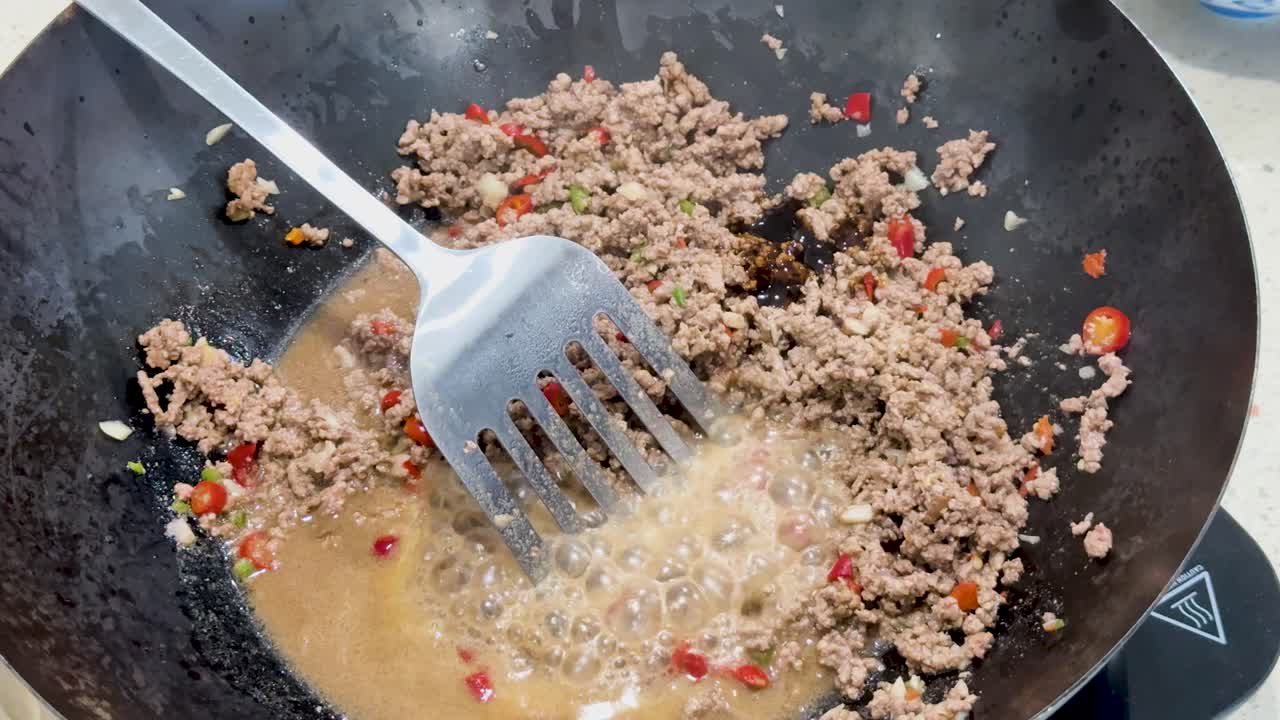 Ground pork is stir fried with sauce and chopped chili in a hot wok under bright lighting, using a metal spatula for mixing