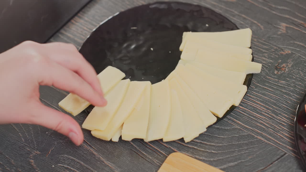 close up of chef using hands to arrange yellow cheese slices in circular pattern on black ceramic plate placed on wooden table, adjusting plate position while organizing dairy pieces with care
