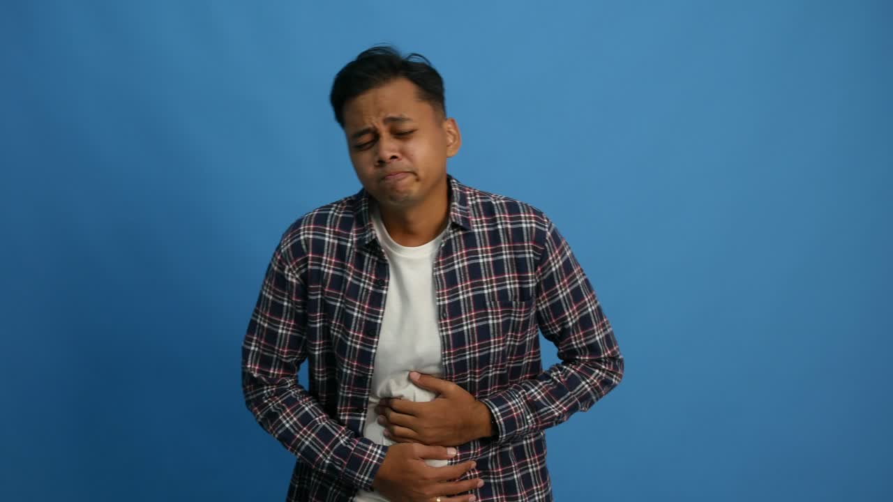 Young Asian man touching his stomach isolated on blue background. Abdominal pain or ache concept