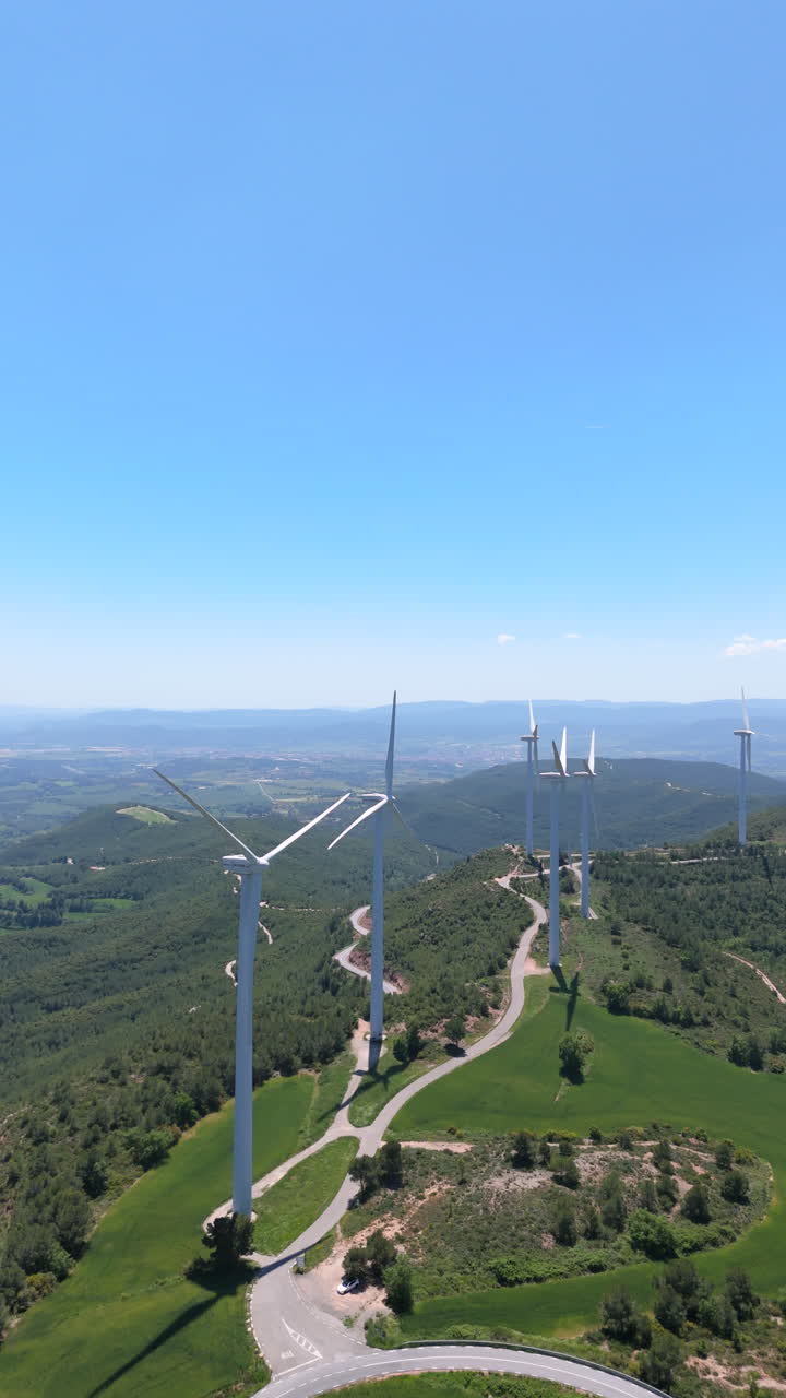 Slow vertical panning shot showing wind turbines on a mountainous landscape with winding roads and green fields. Clean energy infrastructure integrated in a natural environment