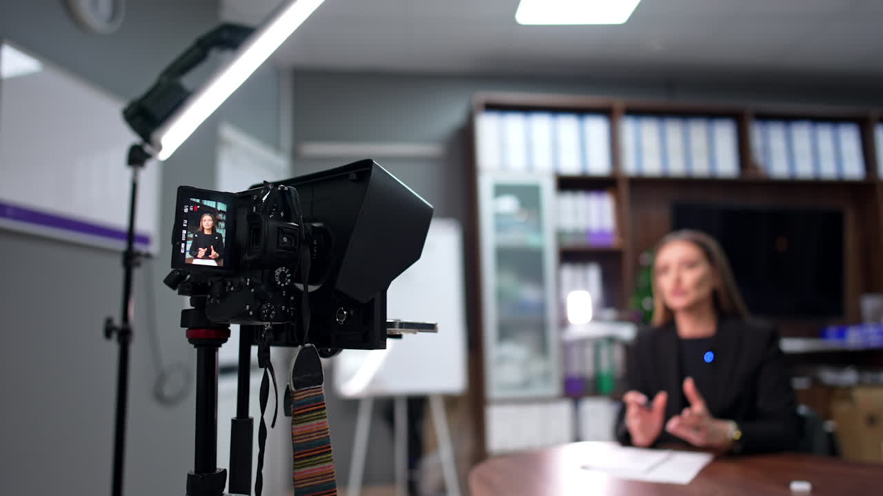 Camera on tripod filming a woman sitting at desk in the office. Recording content for modern blog. Blurred backdrop.
