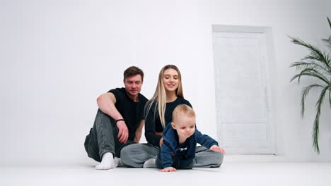 caucasian mom and dad sit on the floor. woman holds an adorable baby who crawls to camera. white backdrop.