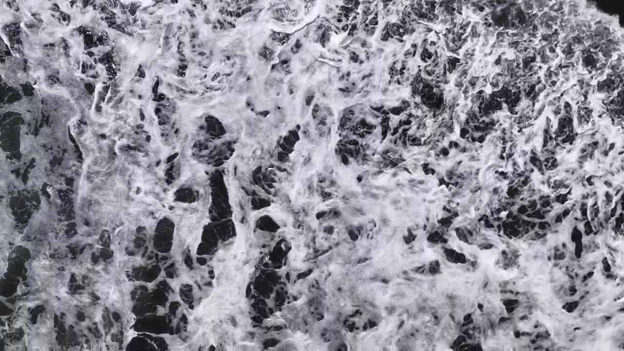 Aerial top-down view of ocean waves breaking along the New Zealand coastline, showing dramatic foam texture.