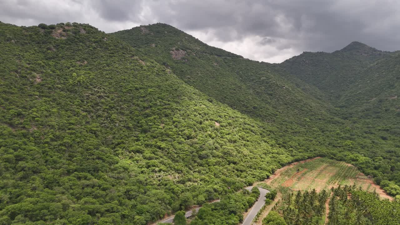 An expansive view of unspoiled nature and towering hills covered in thick green growth. A single paved road highlights a journey through a remote, dramatic mountain pass