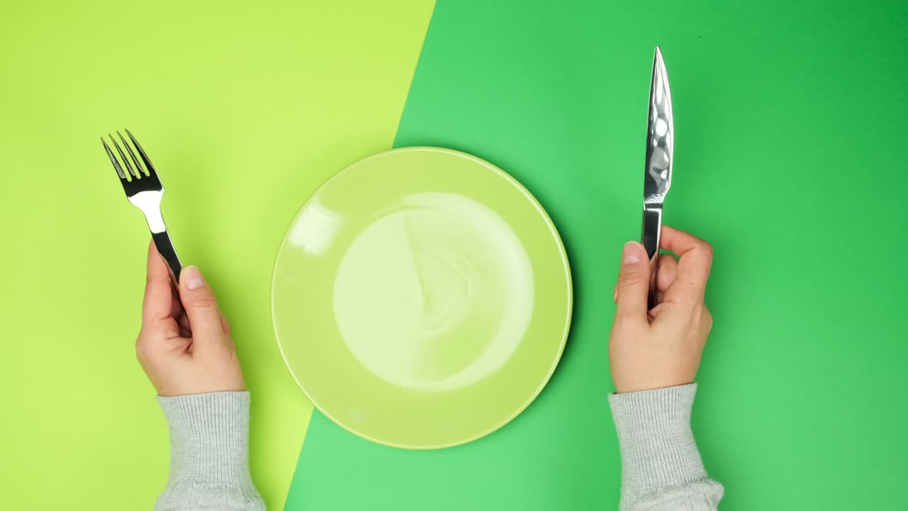 two female hands hold a metal knife and fork, in the middle there is an empty round green plate