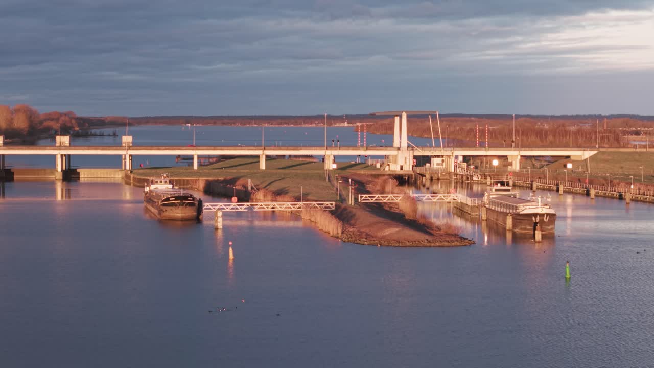 Drone captures canal lock, boats, and bridge surrounded by nature during sunset in a tranquil, scenic setting.