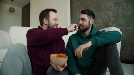 gay couple sitting on the floor having a romantic conversation at home,a man feed his partner with chips