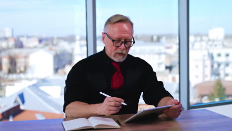confident male businessman wearing black shirt, vest and red tie sits in the office. boss uses i-pad and talks to camera. cityscape in the panoramic window at backdrop.