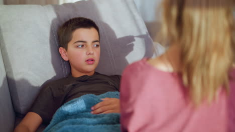 back view of health personnel in maroon shirt collecting thermometer from boy armpit as he lies on couch under blue blanket looking up thoughtfully