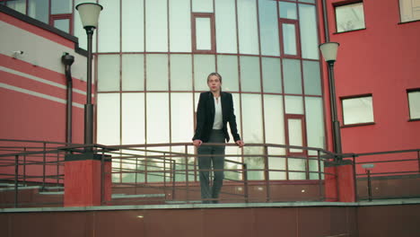 young lady in black blazer rests hands on iron railing while gazing into distance, standing between lamp posts with cctv camera on wall and modern glass building