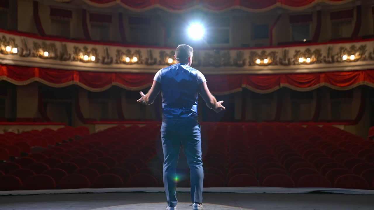 Performer speaks in front of beautiful richly decorated theater hall. Rows of red chairs in theater hall. Big empty theater with one person on stage.