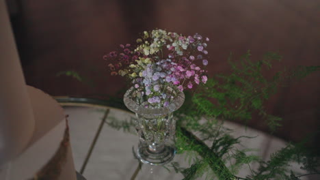 Delicate-bouquet-of-colorful-baby's-breath-flowers-in-a-crystal-vase,-accompanied-by-green-ferns-on-a-reflective-surface