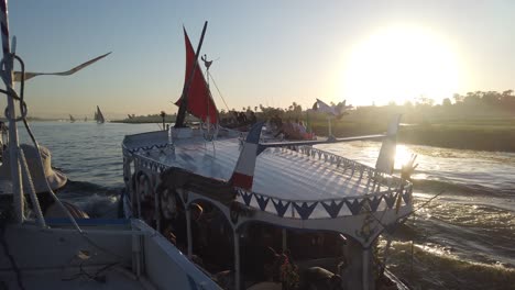 Slow-Motion-of-Multiple-egyption-Local-felucca-tour-boat-on-Nile-river-fully-loaded-with-Tourists-heading-to-Banana-Island-from-Luxor,-Egypt-at-sunset-golden-Hour-passing-by-scenic-fields-on-the-sides