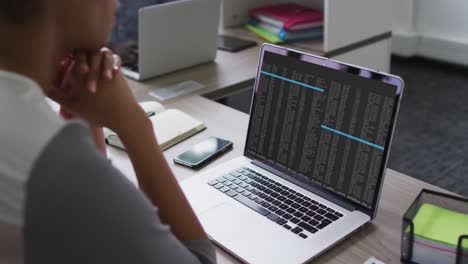 Mixed-race-woman-sitting-at-desk-watching-coding-data-processing-on-laptop-screen